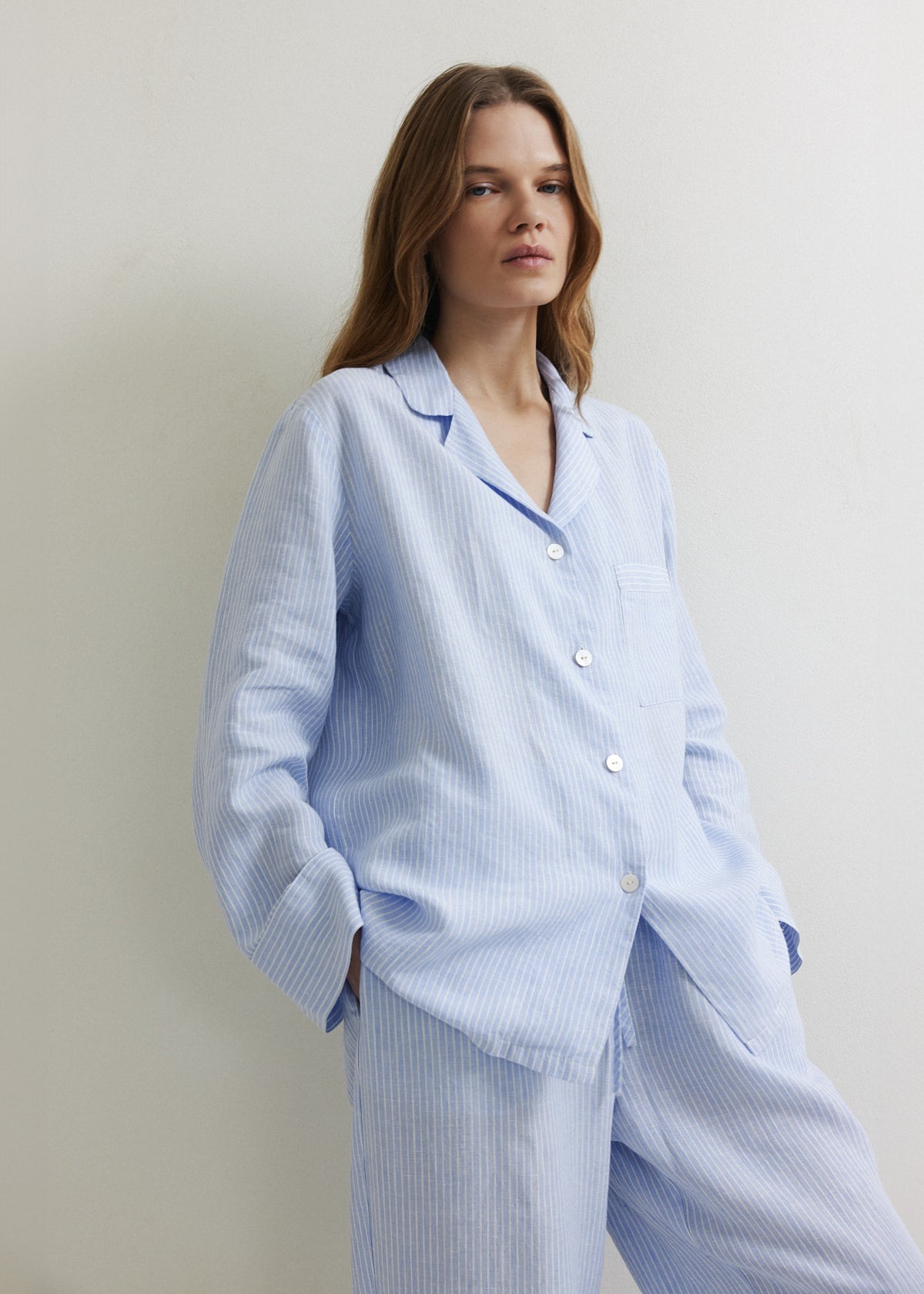 A woman wearing a light blue and white vertically striped sleepwear set, consisting of a long-sleeve button-up shirt and matching trousers, standing against a plain off-white wall.