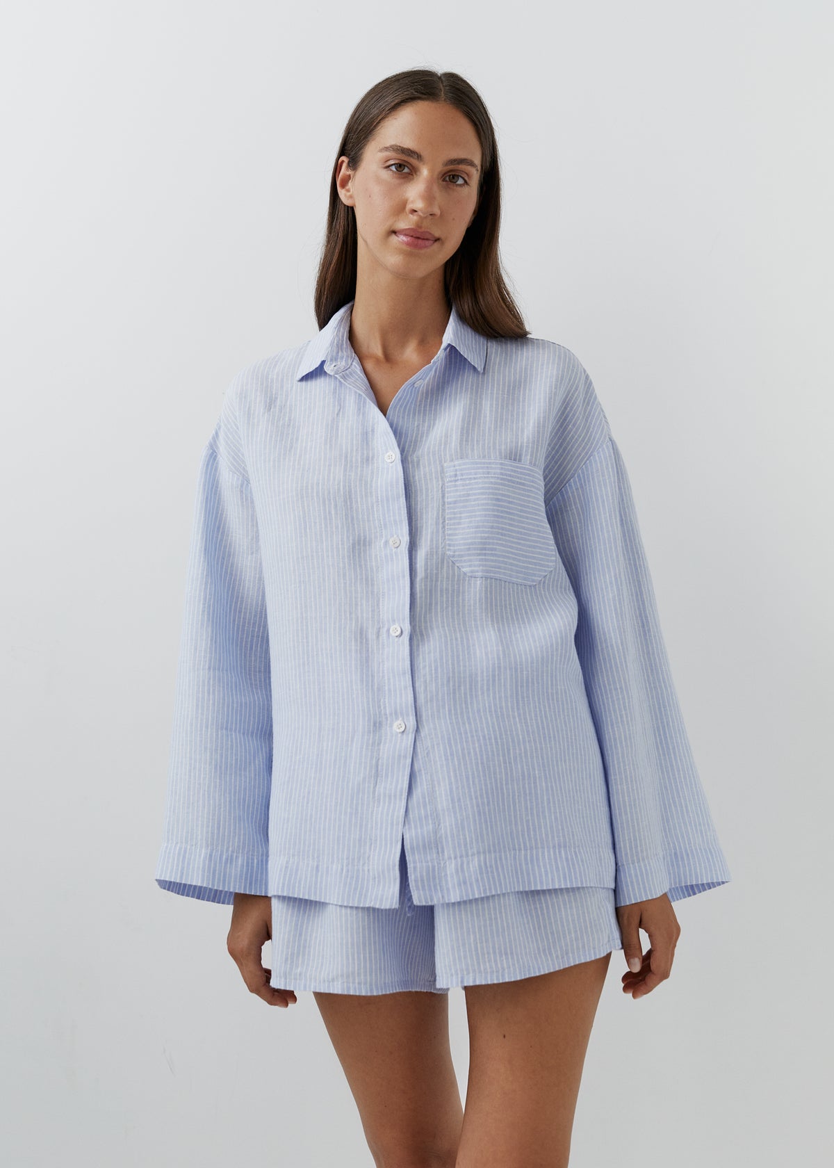 A woman modeling a light blue and white vertically striped button-up shirt and matching shorts set against a plain light gray studio background.