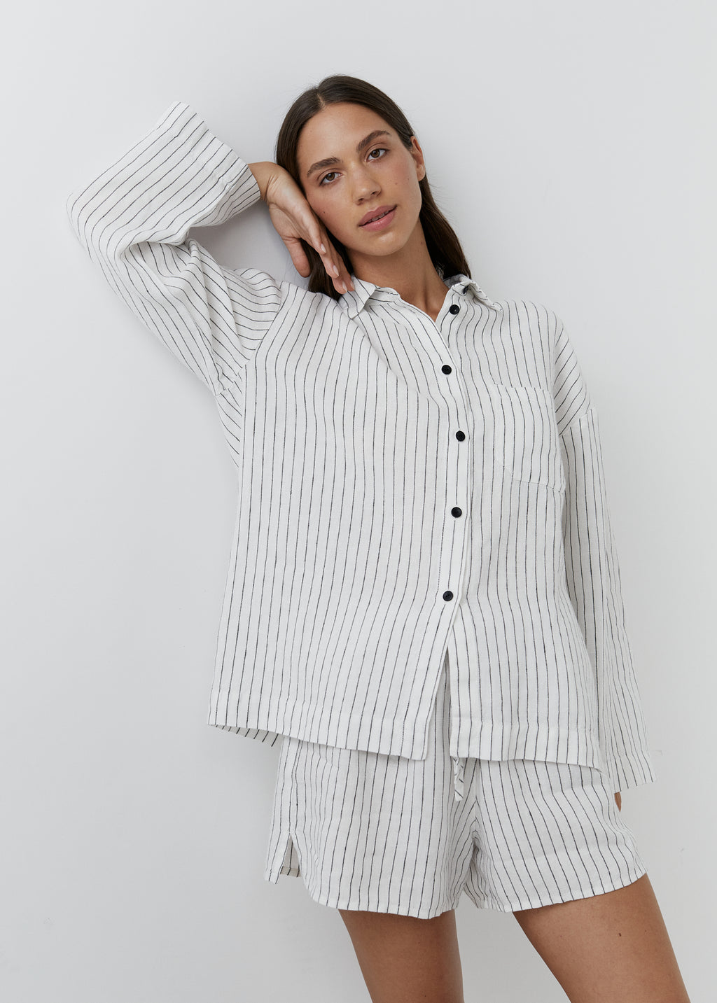A woman wearing a matching two-piece pajama set with a white base and thin black vertical pinstripes, consisting of a long-sleeve button-up shirt and coordinating shorts, posed against a light gray studio background.