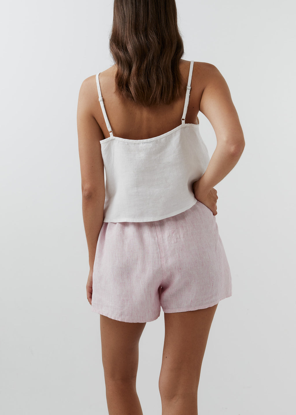 A woman shown from the back wearing a white camisole top and light striped pink linen shorts against a plain light gray studio background.