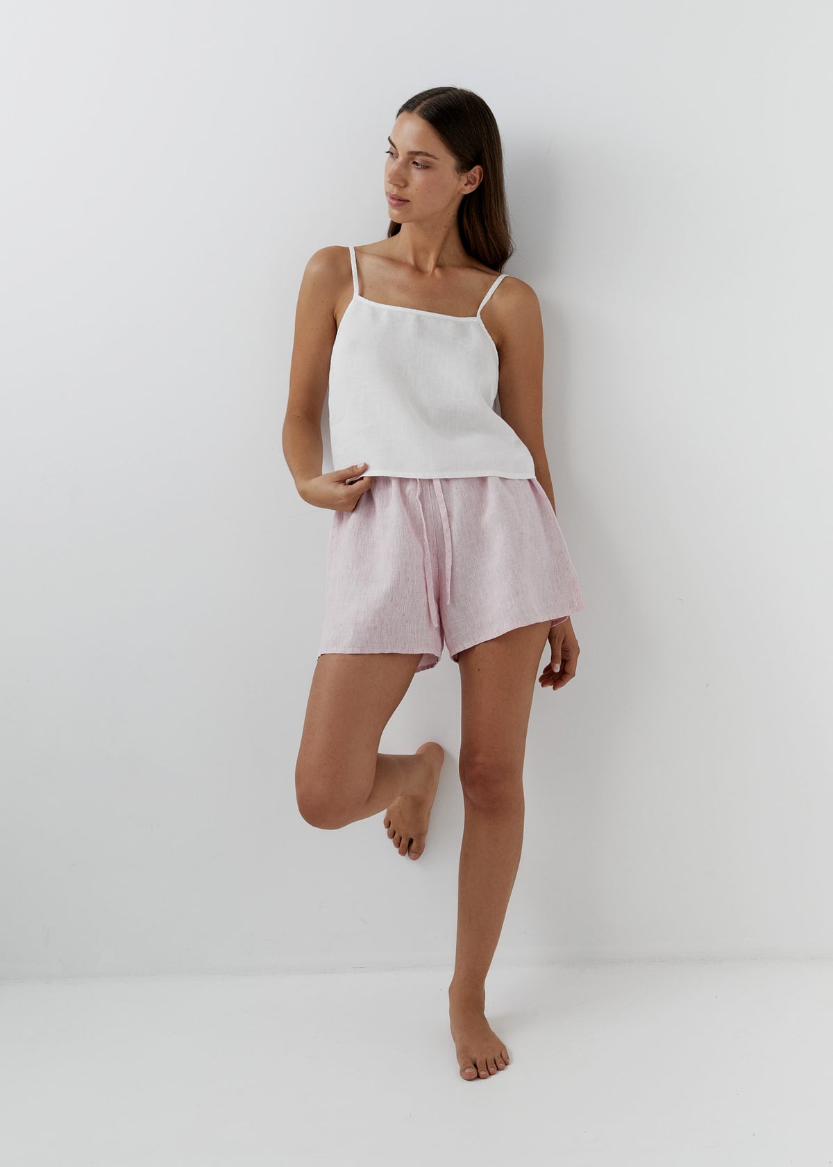 A woman modeling a two-piece loungewear set consisting of a white camisole top and striped light pink linen shorts, standing against a plain white wall.