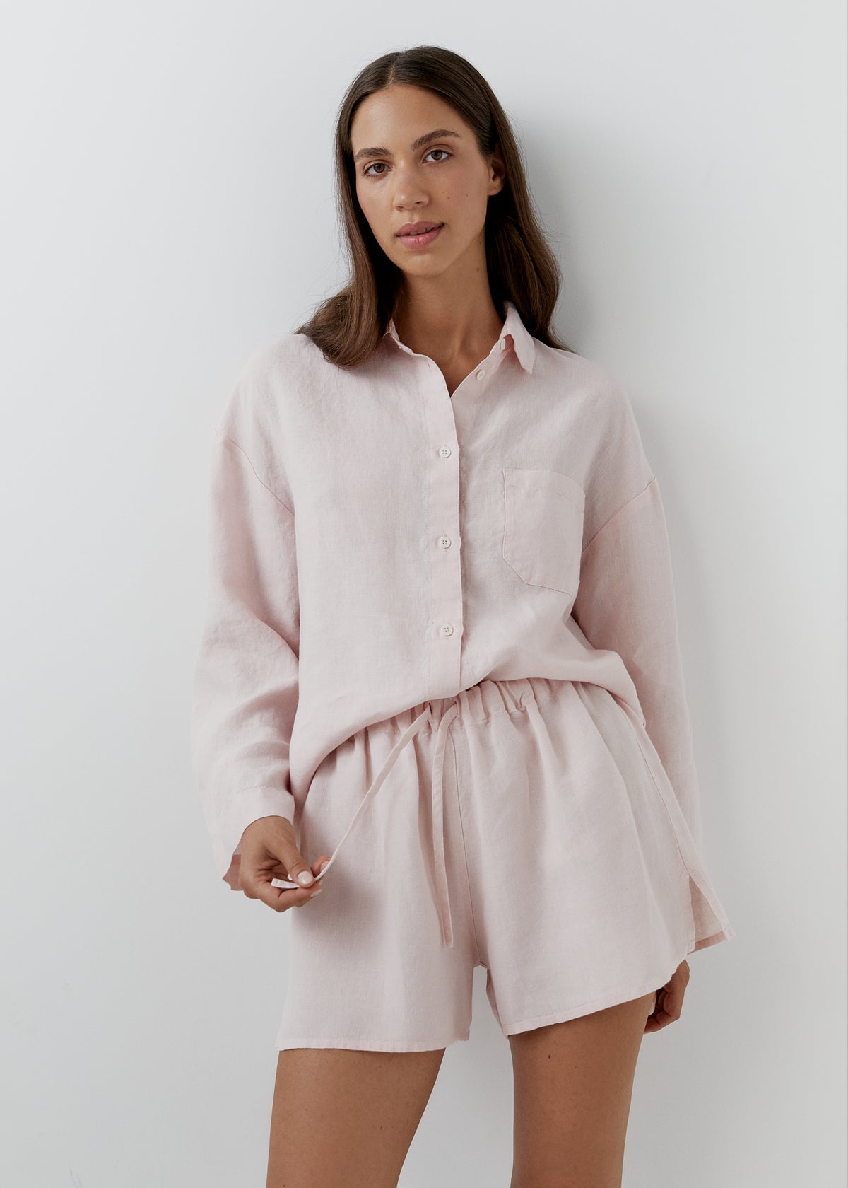 A woman wearing a light pink linen button-up shirt and matching drawstring shorts, standing against a plain light gray wall.