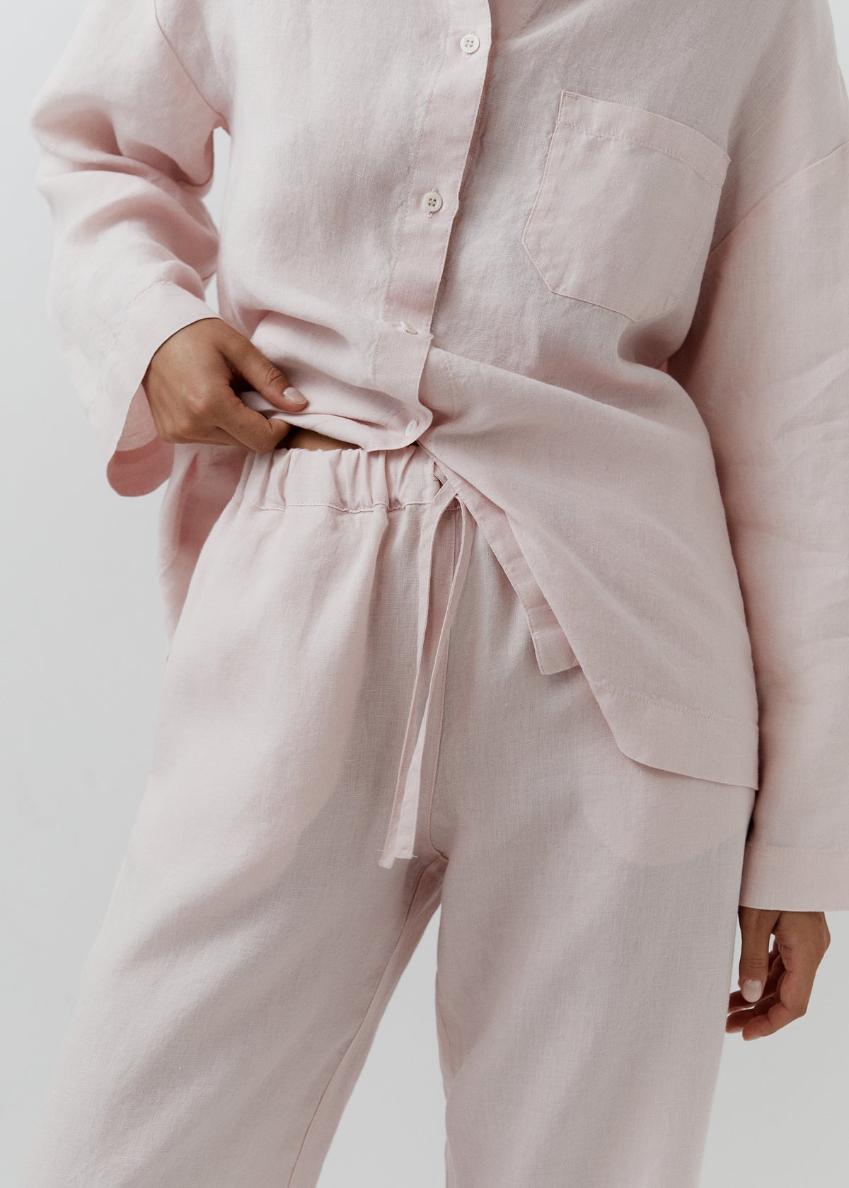 A close-up of a person wearing a light blush pink linen pajama set, featuring a long-sleeve button-up shirt and matching drawstring pants, against a plain light gray background.