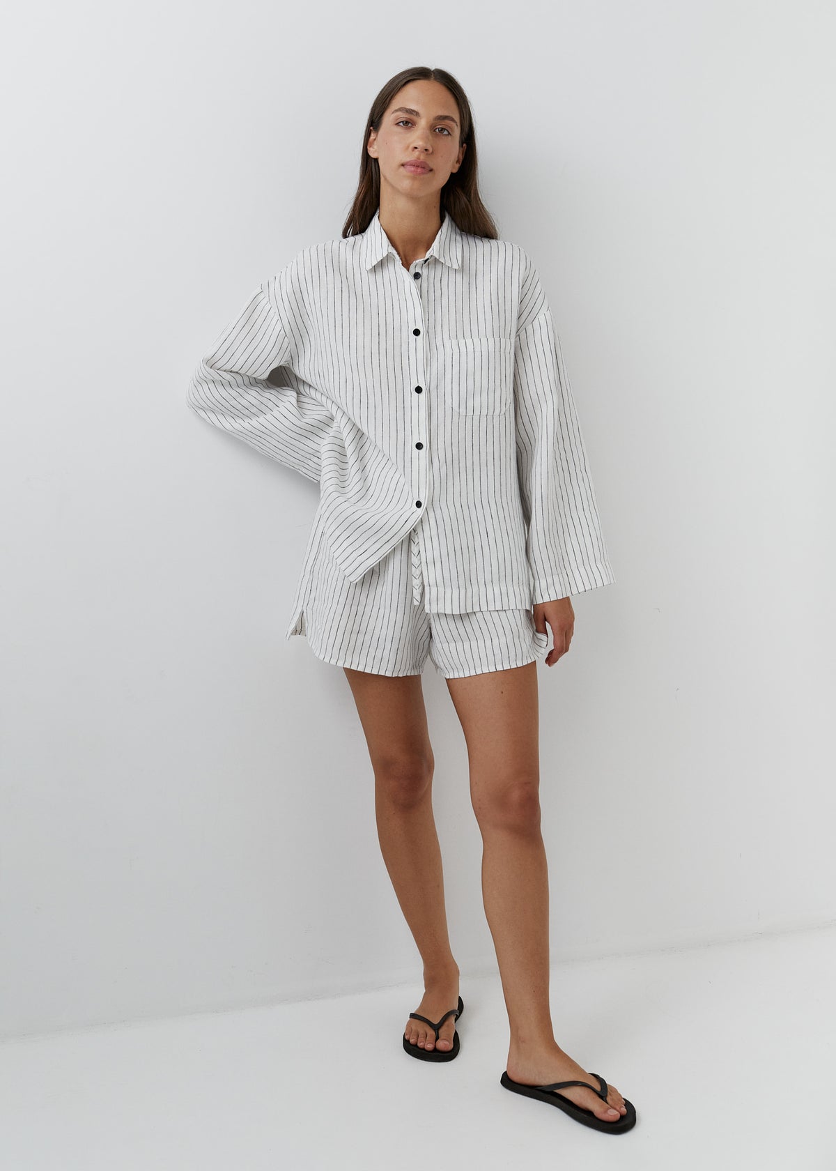 A woman modeling a matching two-piece pajama set consisting of a long-sleeve button-up shirt and shorts, both in a white and gray vertical stripe pattern, paired with black flip-flops against a plain white studio background.