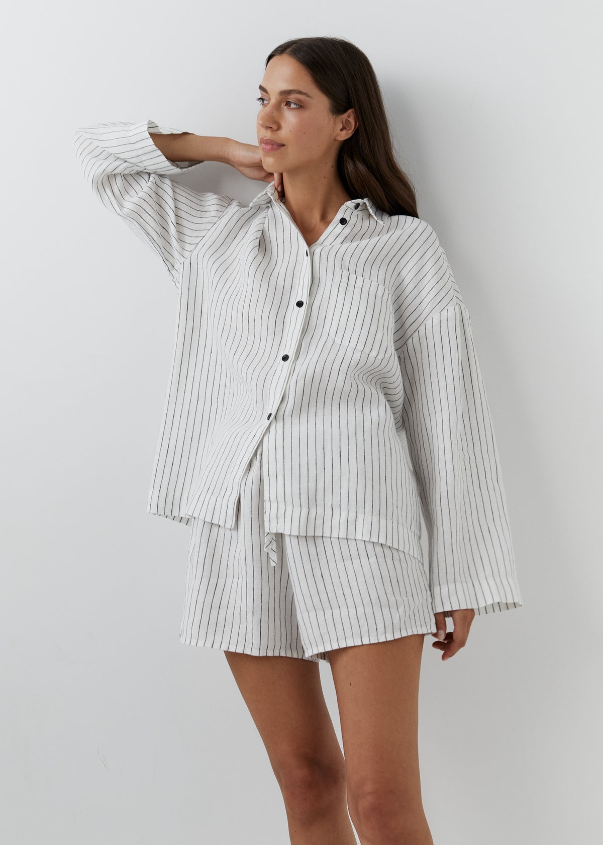 A woman modeling a matching two-piece pajama set consisting of a long-sleeve button-up shirt and shorts, both featuring a white base with thin vertical black pinstripes, set against a plain light gray wall.
