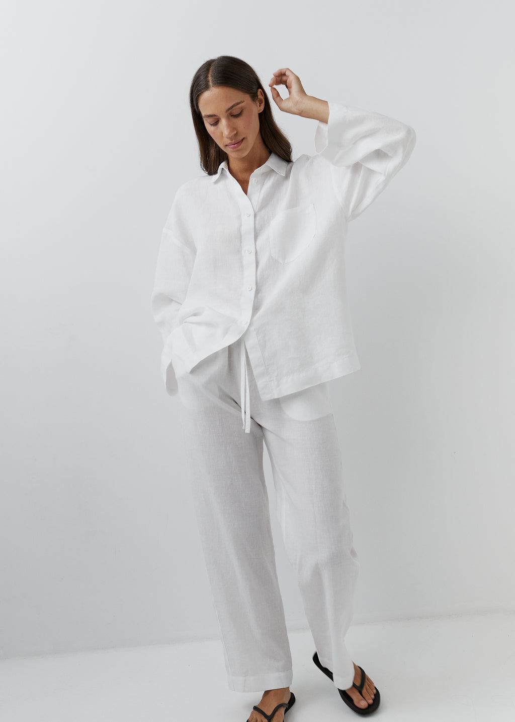 A woman modeling a white linen pyjama set consisting of a long-sleeve button-up shirt and matching wide-leg pants, styled with black sandals against a clean white studio background.