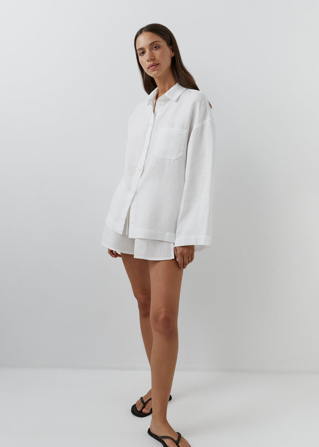 A woman modeling a white linen button-up shirt and matching shorts set against a plain white studio background.