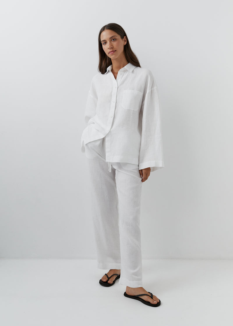 A woman modeling a white two-piece lounge set consisting of a long-sleeve button-up shirt and matching wide-leg pants, paired with black flip-flop sandals, set against a plain white studio background.