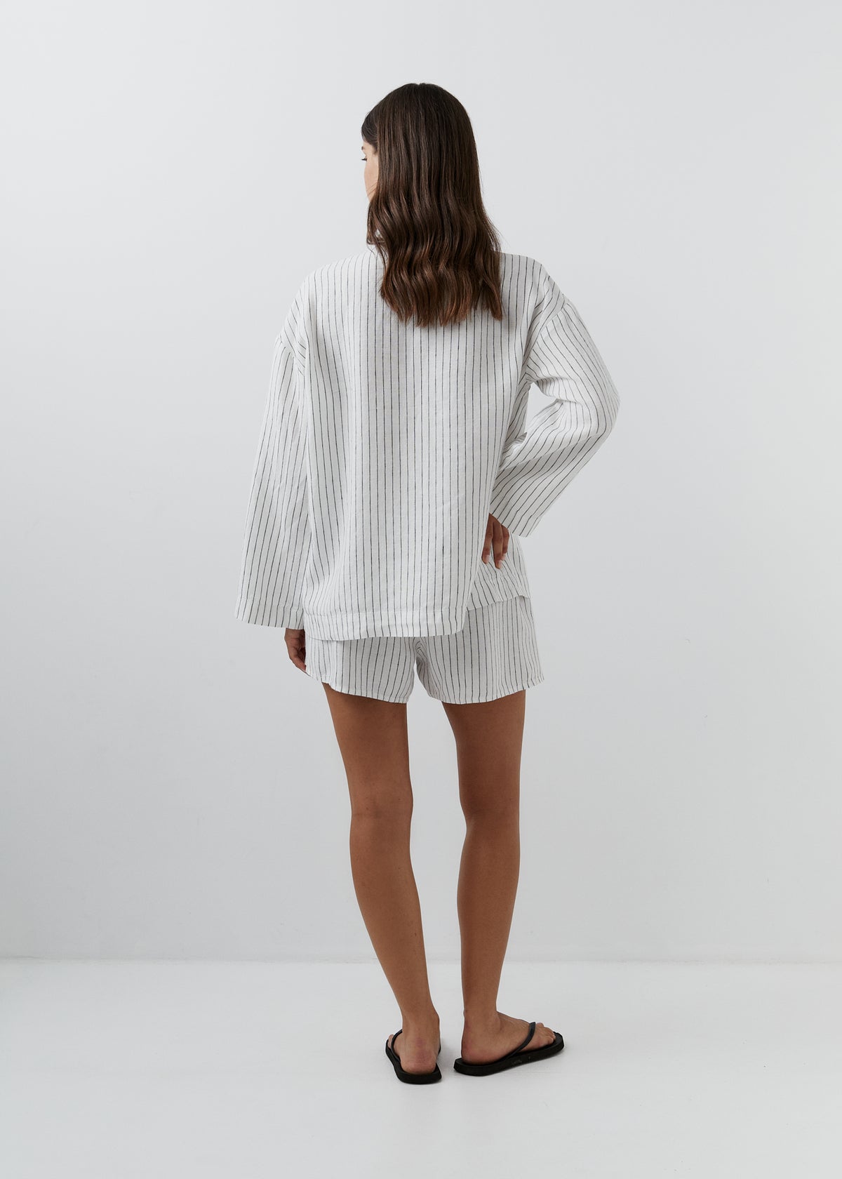 A woman viewed from behind wearing a matching white and gray vertically striped lounge set consisting of a long-sleeve top and shorts, paired with black flip-flops against a plain white studio background.