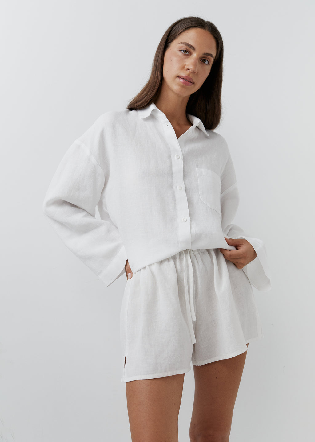 A woman modeling a white long-sleeve button-up shirt and matching white shorts set against a plain light gray studio background.