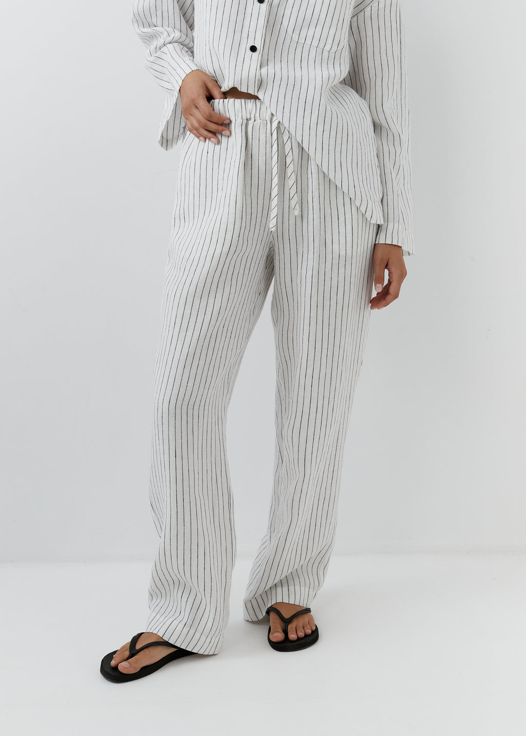 A person wearing a matching white and black vertically striped pajama set with a drawstring waist, shown from the waist down against a plain light gray studio background.