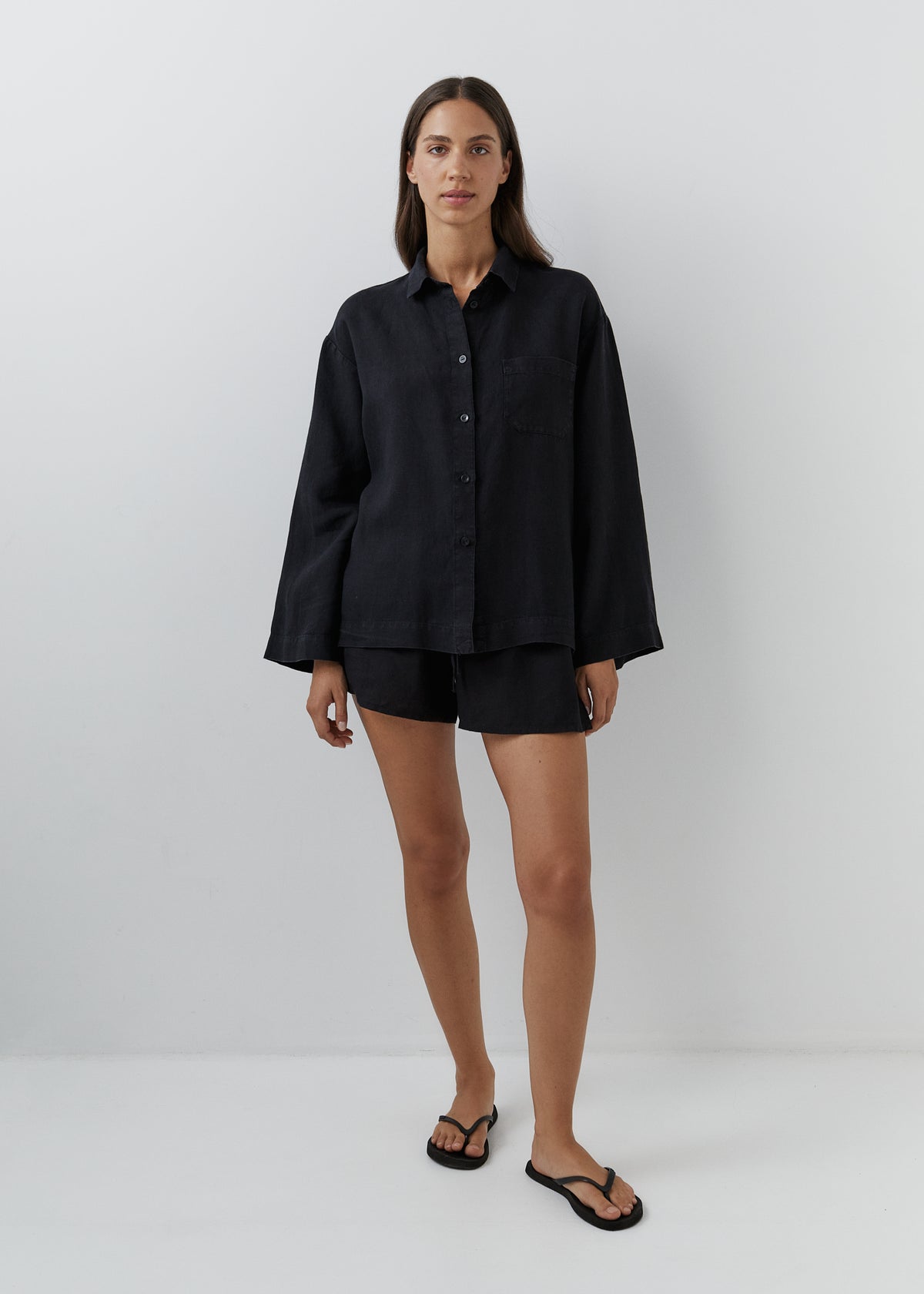 A woman modeling a black long-sleeve button-up shirt and matching black shorts against a plain light gray studio background.
