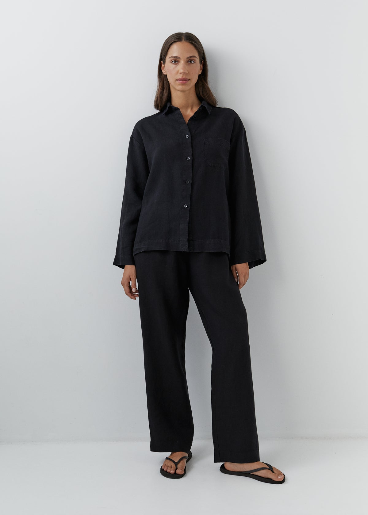 A woman stands against a plain light gray studio wall wearing a loose black long-sleeve button-front shirt with matching black wide-leg pants and black sandals.