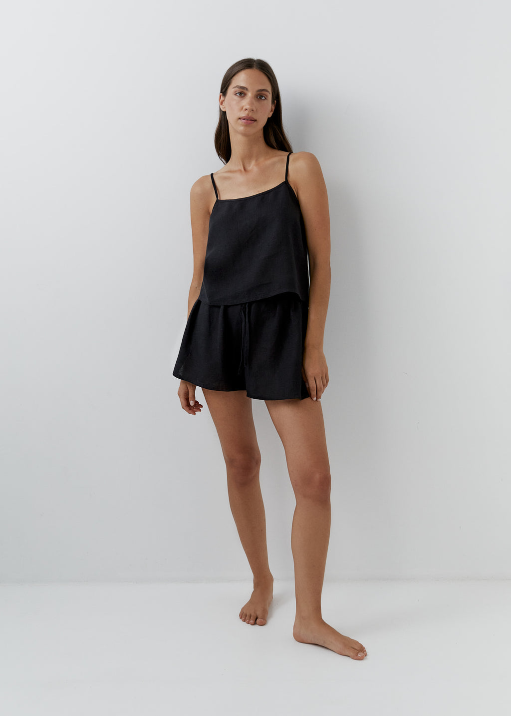 A woman stands barefoot against a plain light gray studio wall wearing a black camisole top and matching black shorts.