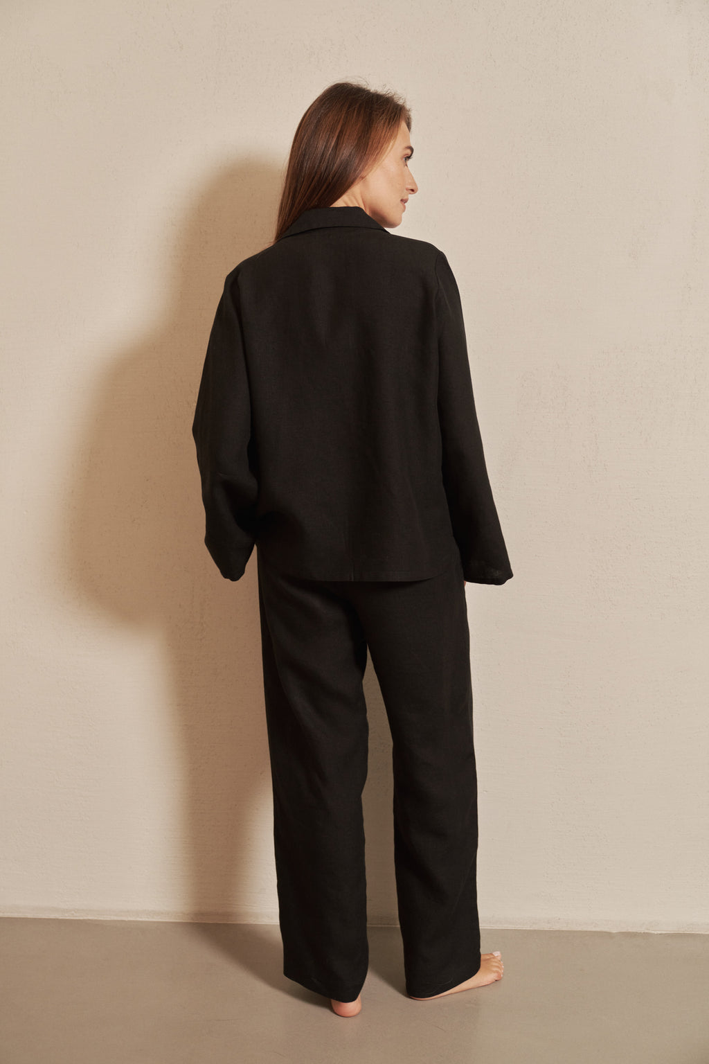 A woman stands with her back to the camera wearing a black long-sleeve blazer and matching wide-leg trousers against a plain beige wall.