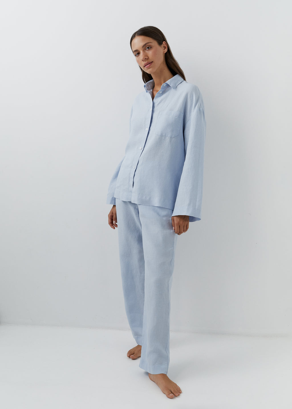 A woman modeling a light blue two-piece pyjama set consisting of a long-sleeve button-up shirt and matching wide-leg pants, standing against a plain white studio background.