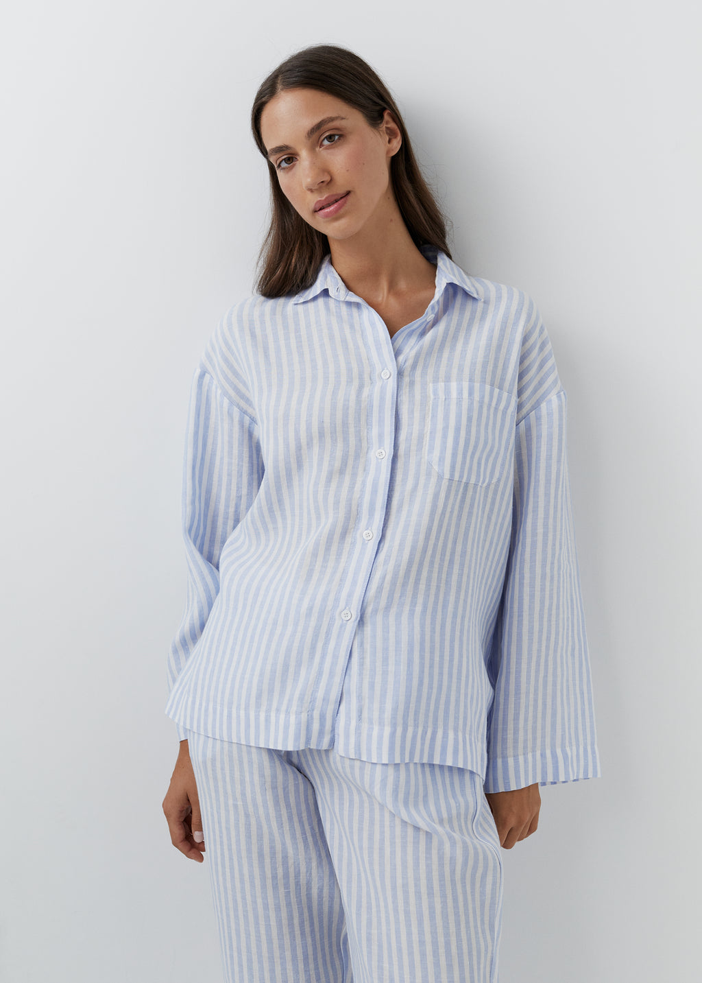 A woman modeling a light blue and white vertically striped long-sleeve button-up shirt and matching striped pajama pants against a plain light gray studio background.