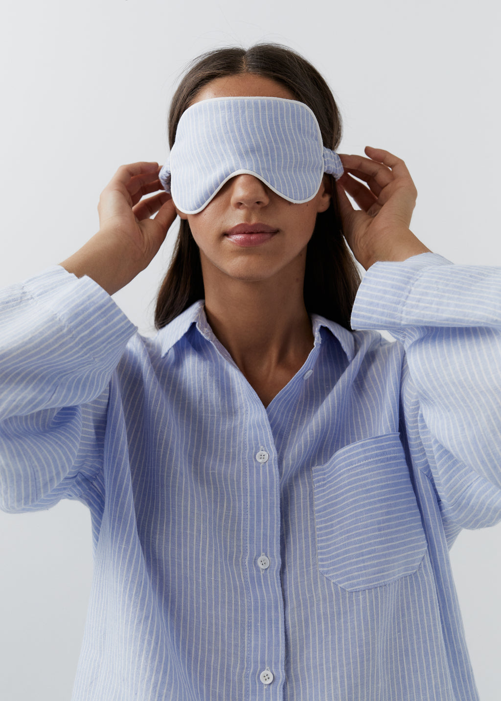 A woman wearing a light blue and white striped sleep mask and a matching long-sleeve button-up shirt, adjusting the mask with both hands against a plain light gray background.