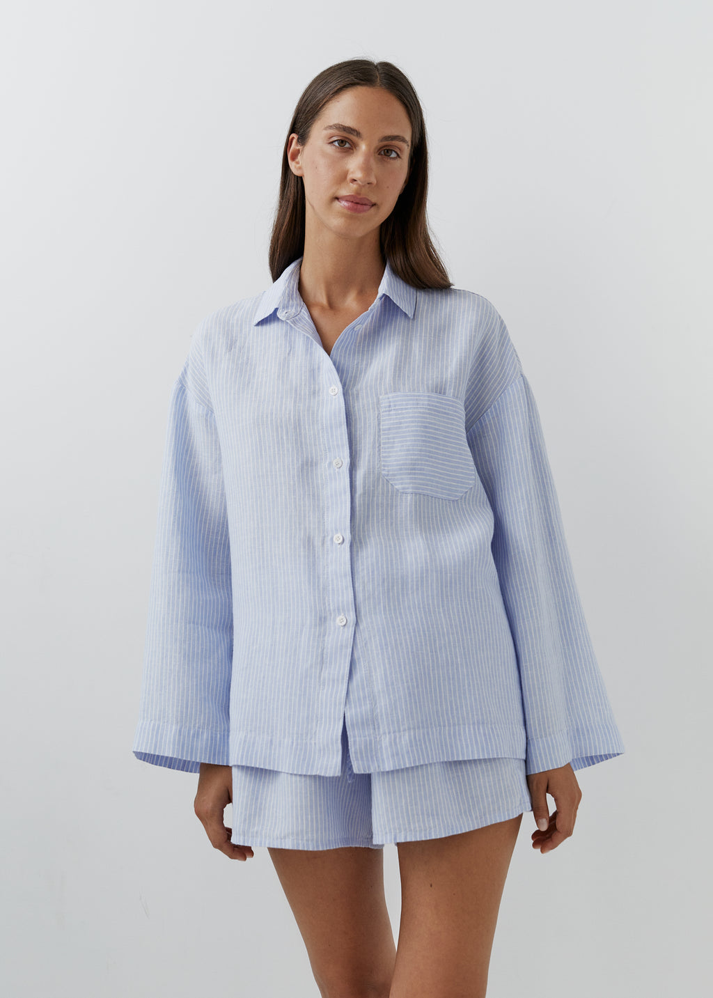 A woman modeling a light blue and white vertically striped button-up shirt and matching shorts set against a plain light gray studio background.