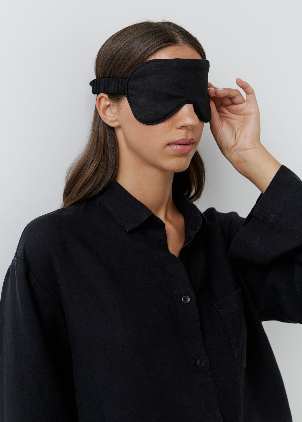 A woman wearing a black sleep mask and a black button-up shirt, adjusting the strap of the mask against a plain light gray background.