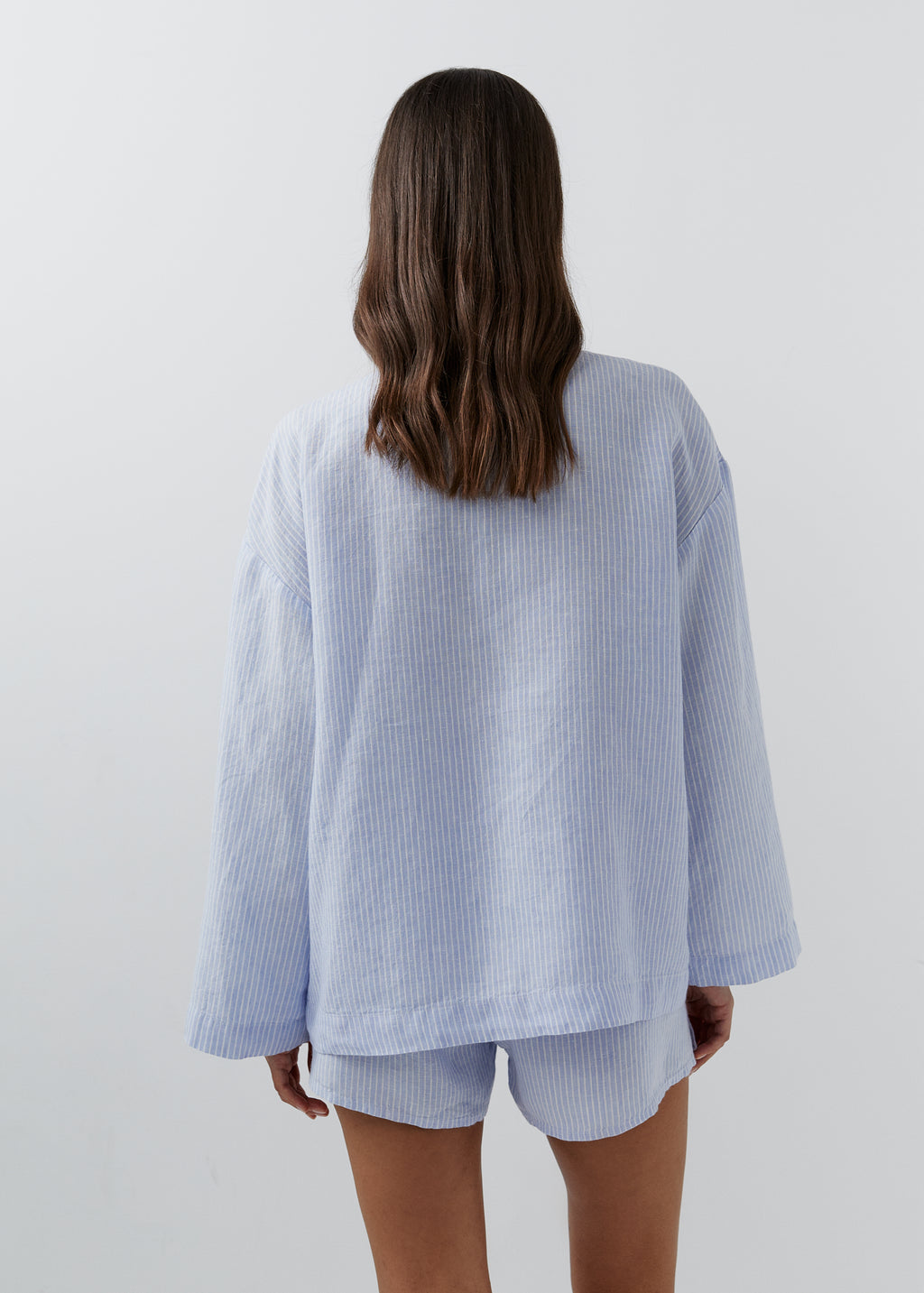 A woman viewed from behind wearing a light blue and white vertically striped long-sleeve top and matching shorts against a plain light gray background.