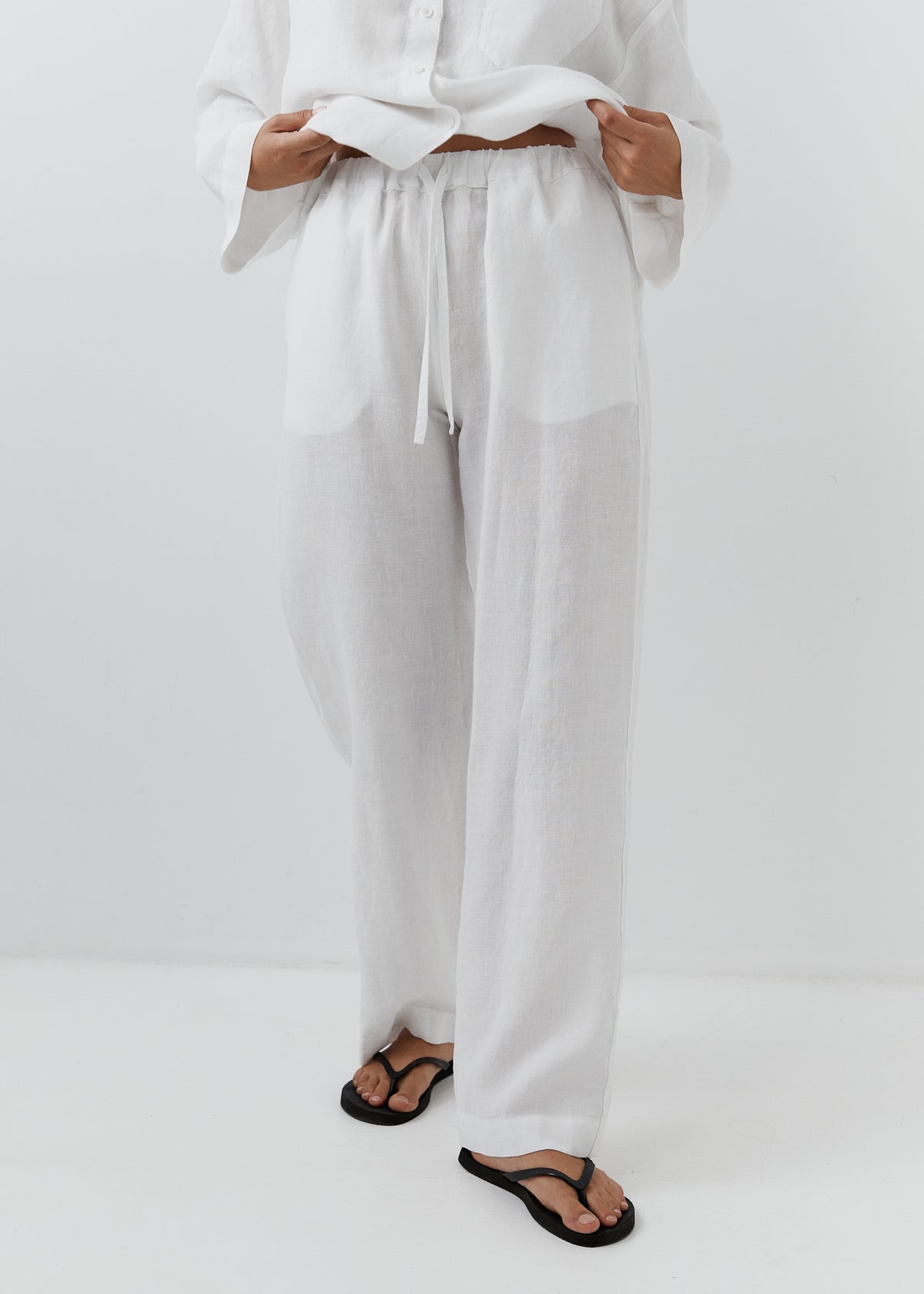 White wide-leg linen pants with a drawstring waist, shown on a model wearing black flip-flops against a white studio background.
