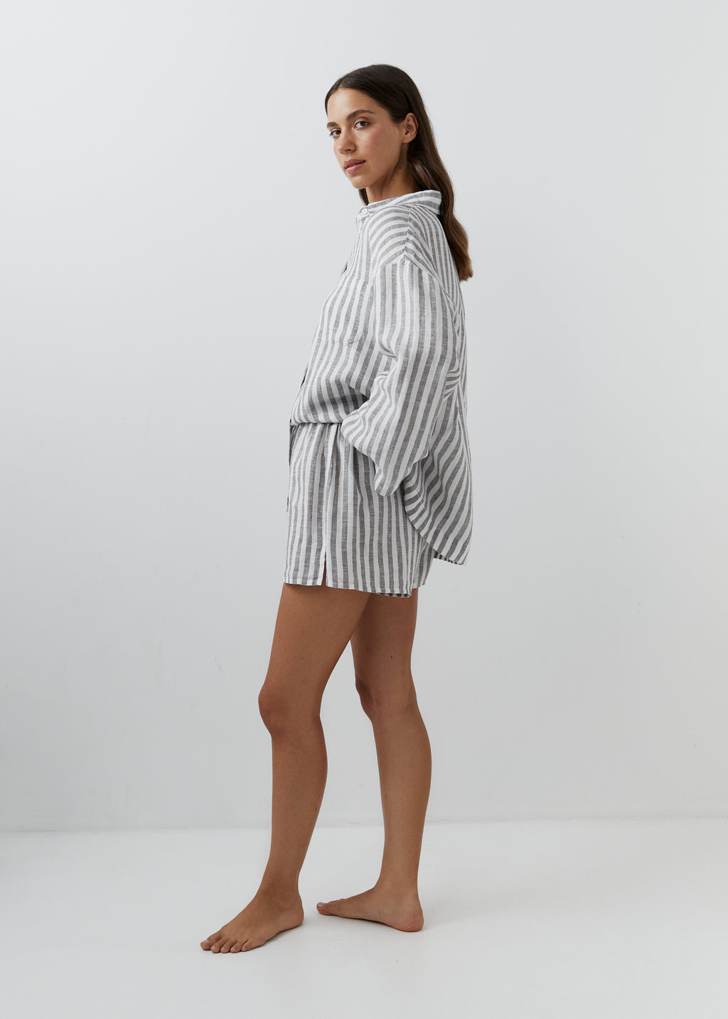 A woman modeling a matching striped lounge set consisting of a long-sleeve top and shorts, featuring vertical gray and white stripes, photographed against a plain white studio background.