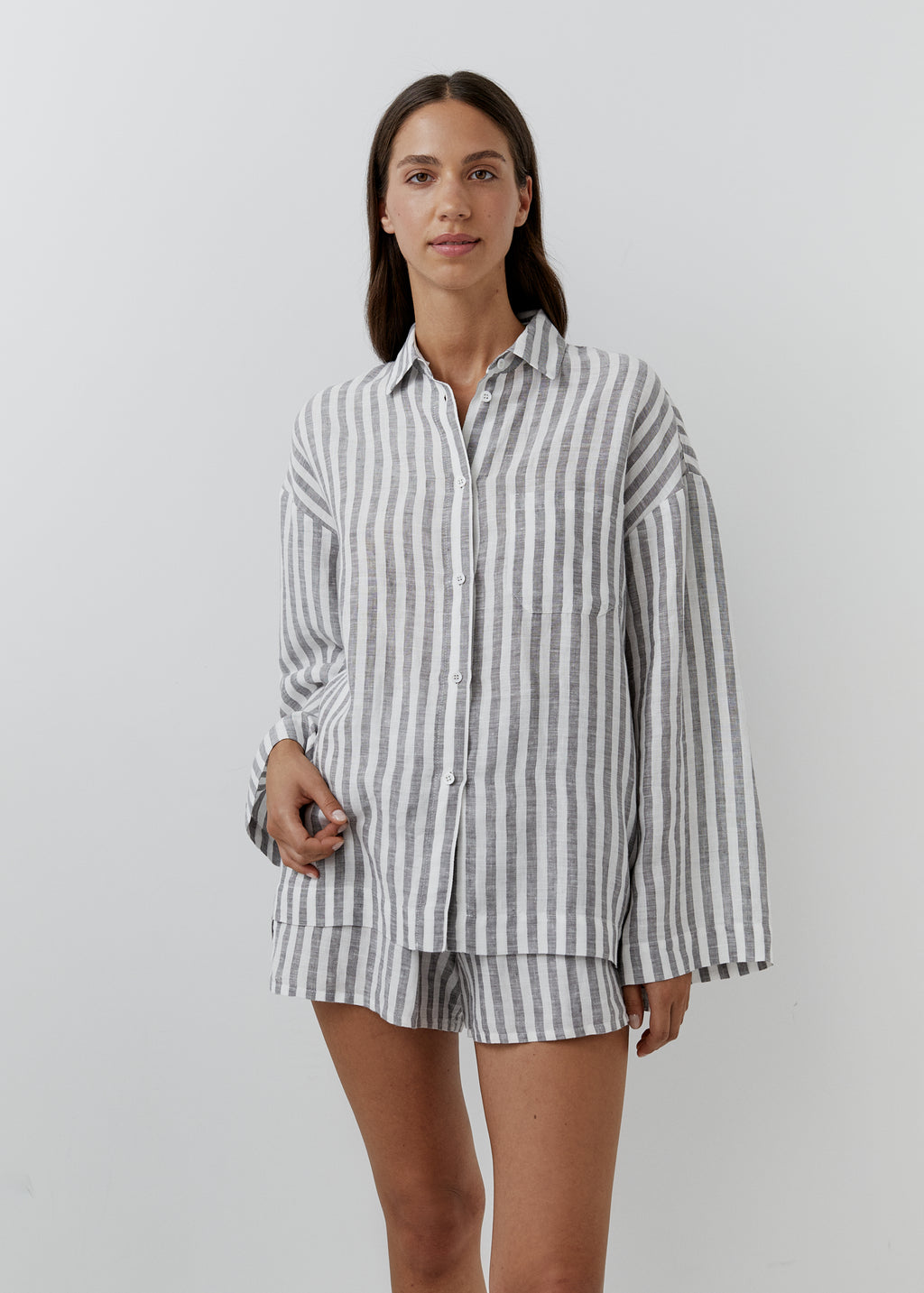 A woman modeling a matching long-sleeve button-up shirt and shorts set in a gray and white vertical stripe linen fabric, shot against a plain light gray studio background.