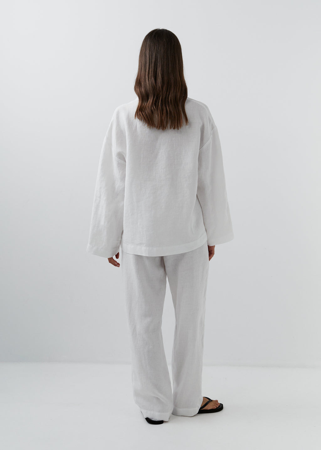 A woman viewed from behind wearing a loose white linen two-piece set consisting of a long-sleeve top and wide-leg trousers, paired with black sandals, against a plain white studio background.