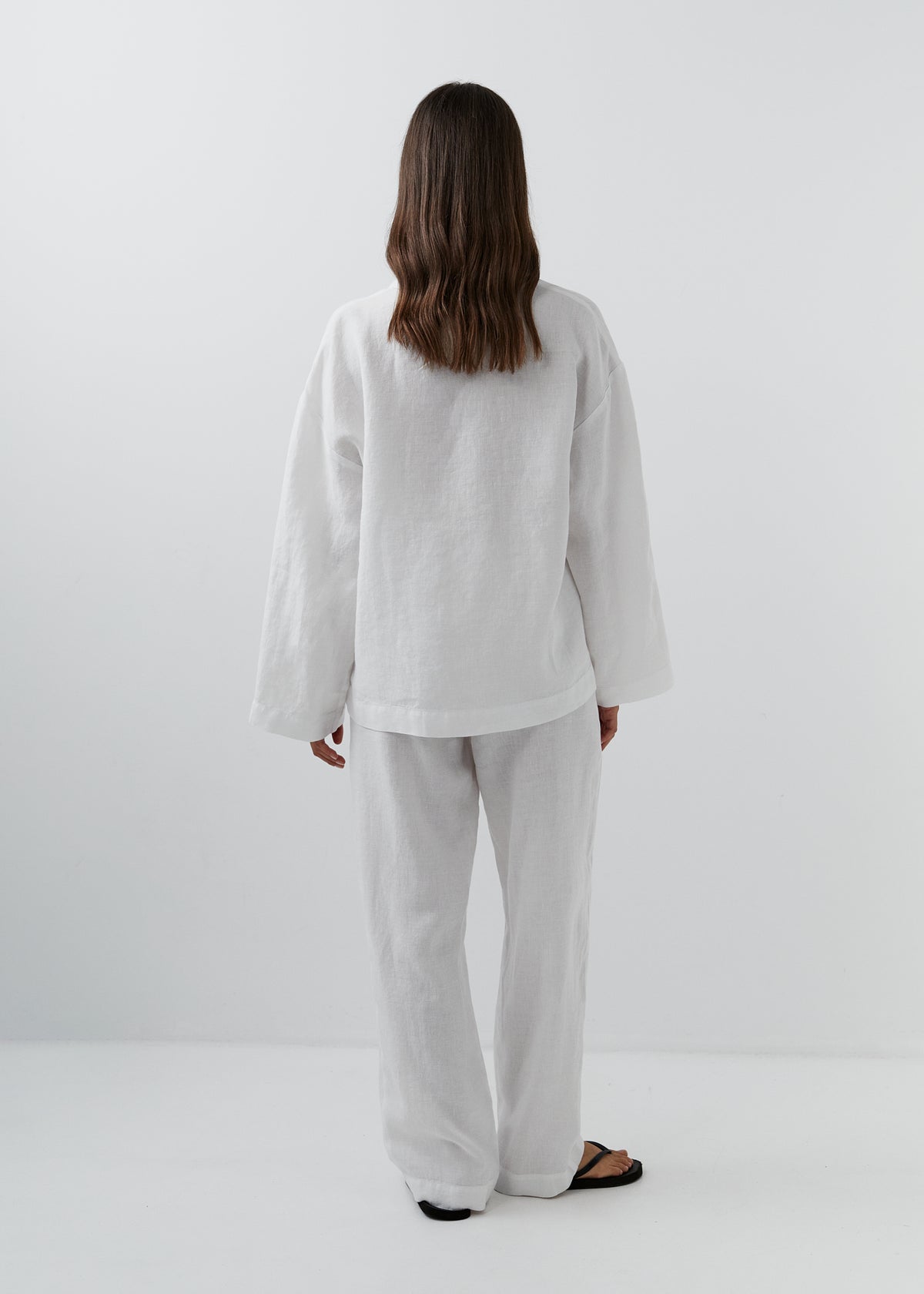 A woman viewed from behind wearing a loose white linen two-piece set consisting of a long-sleeve top and wide-leg trousers, paired with black sandals, against a plain white studio background.