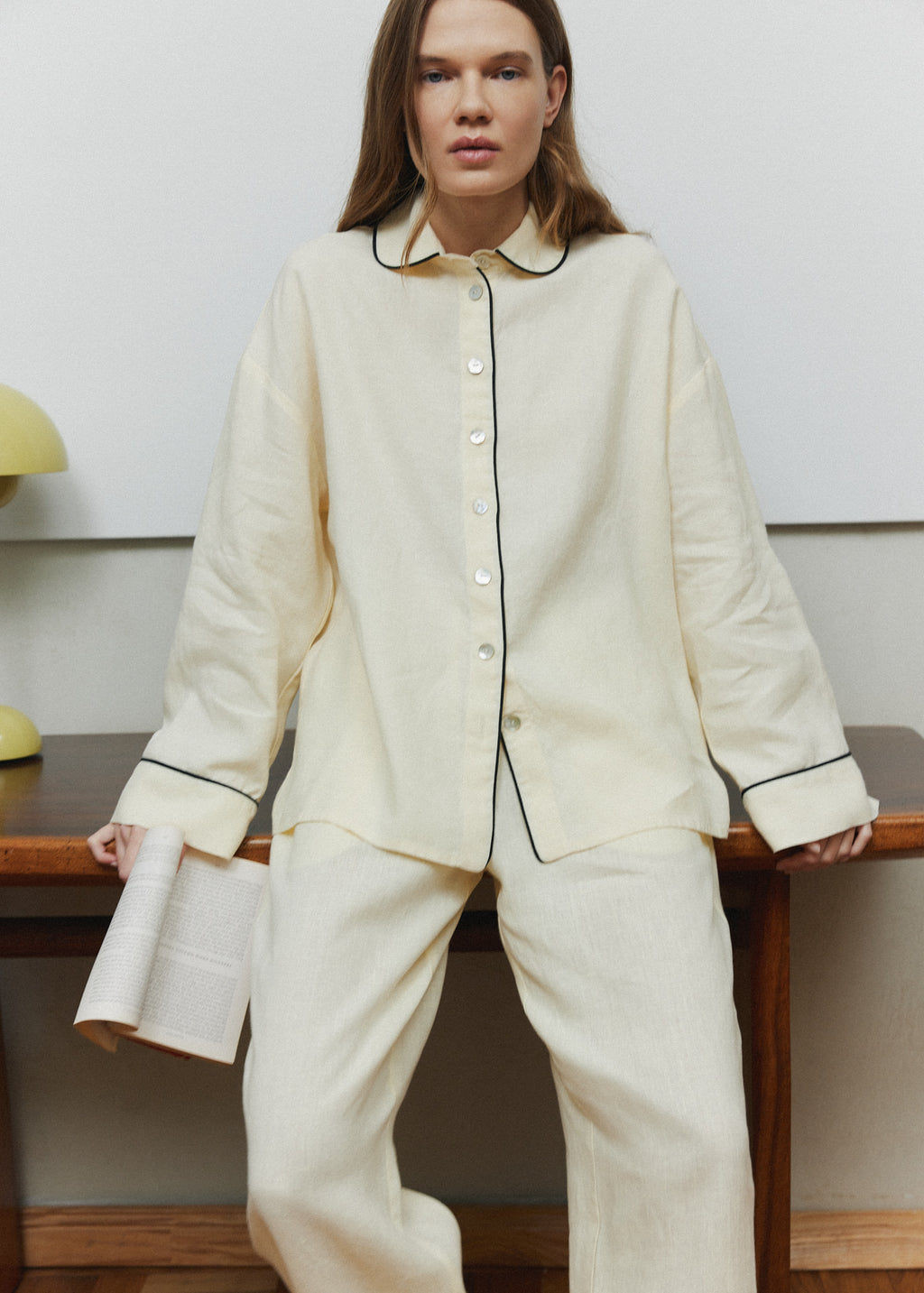 Pastel yellow linen pajama set with thin black piping on the collar, cuffs and placket, button-up shirt and relaxed trousers, worn at home.