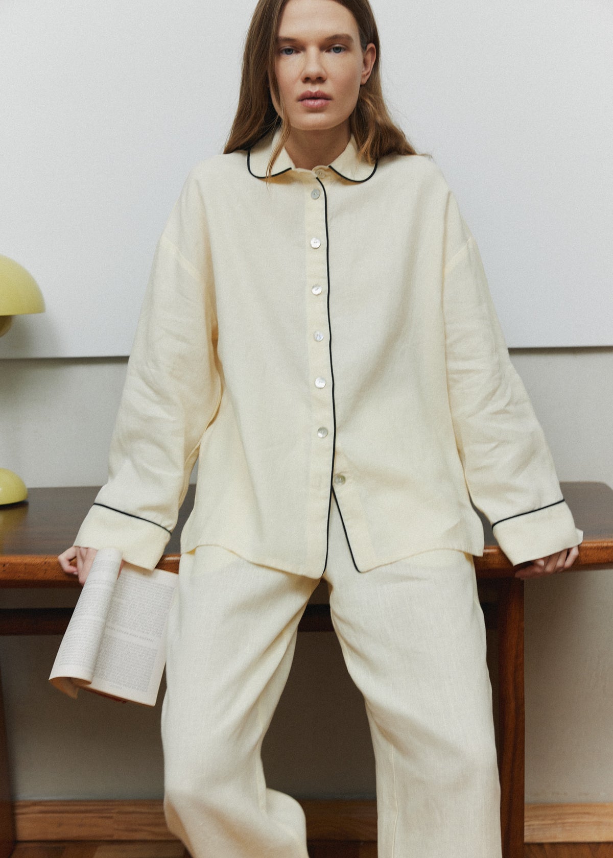 Pastel yellow linen pajama set with thin black piping on the collar, cuffs and placket, button-up shirt and relaxed trousers, worn at home.