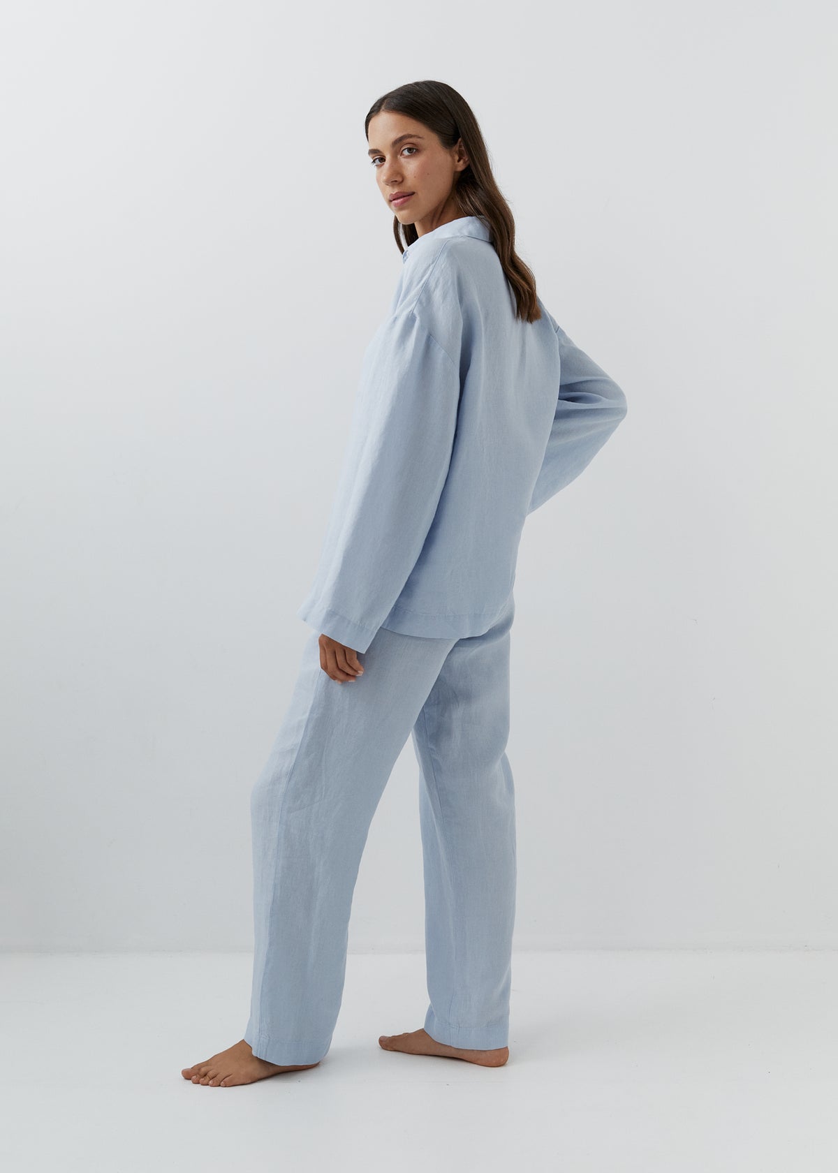 A woman modeling a light blue two-piece pajama set consisting of a long-sleeve button-up top and matching wide-leg pants, standing barefoot against a plain white studio background.