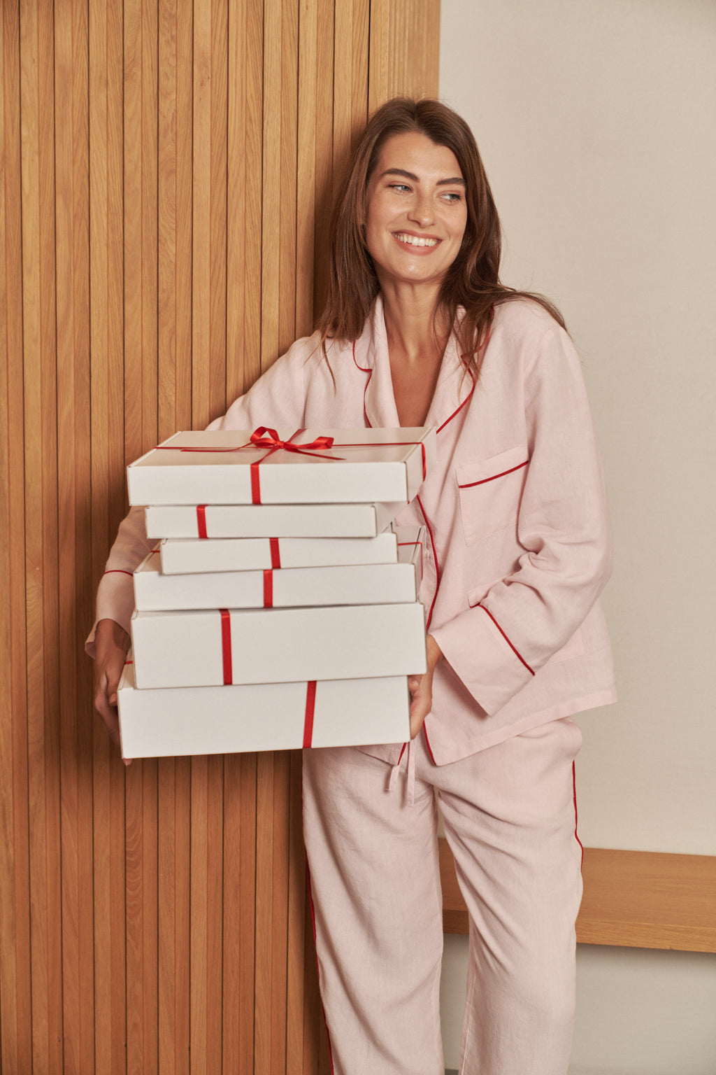 Light pink linen pajama set with red piping, worn by a woman holding white Christmas gift boxes tied with red ribbon, a linen pajama gift for her.