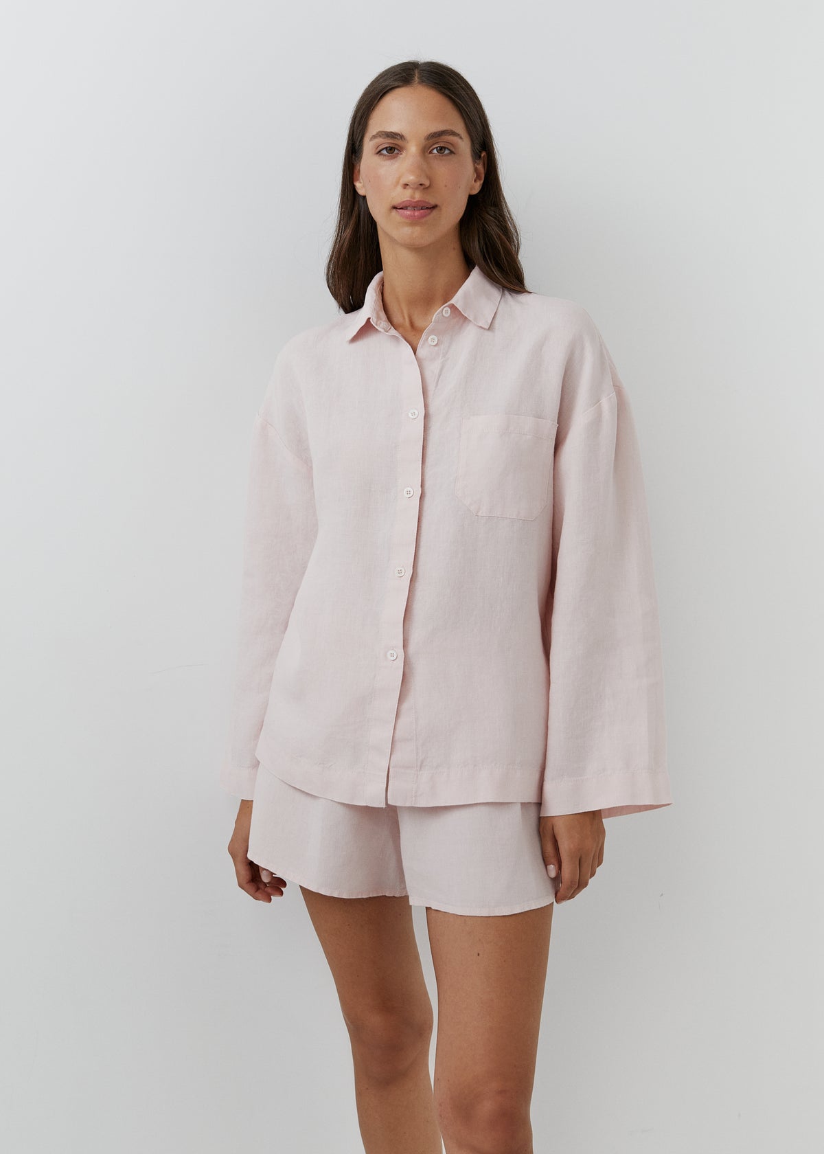 A woman modeling a light pink long-sleeve button-up shirt and matching shorts set against a plain light gray studio background.
