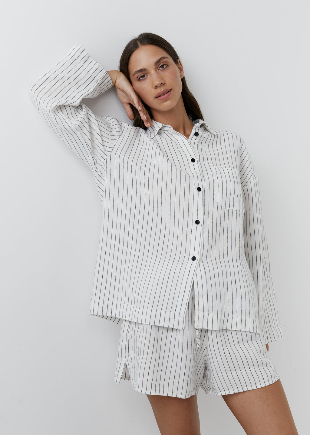 A woman wearing a matching two-piece pajama set with a white base and thin black vertical pinstripes, consisting of a long-sleeve button-up shirt and coordinating shorts, posed against a light gray studio background.