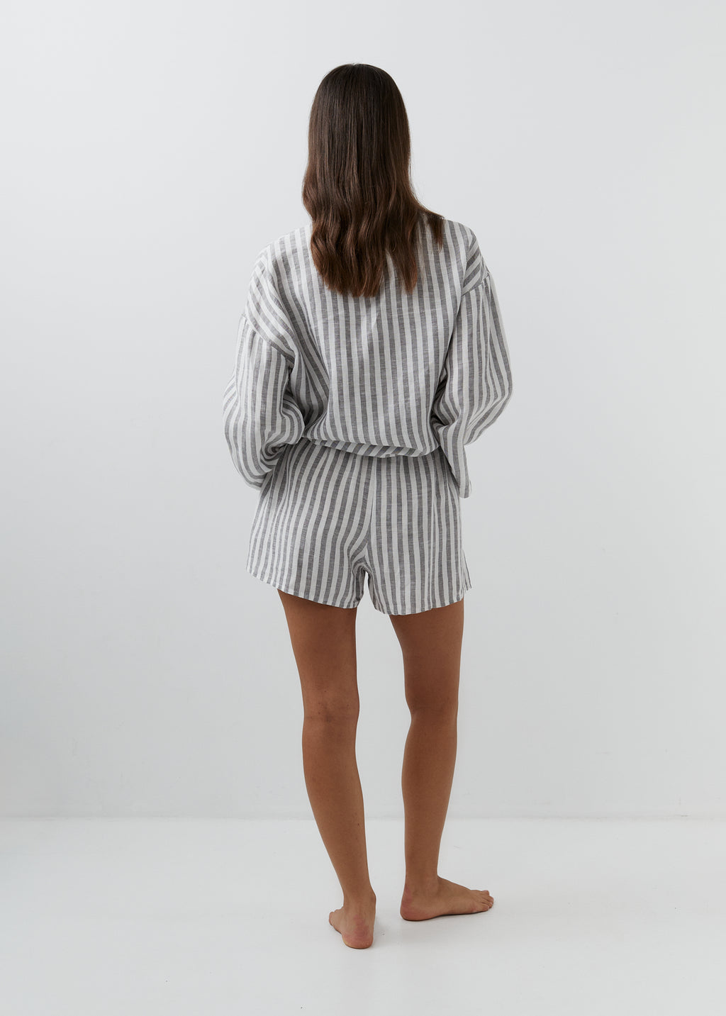 A woman viewed from behind wearing a matching gray and white vertically striped lounge set consisting of a long-sleeve top and shorts, standing barefoot against a plain white studio background.