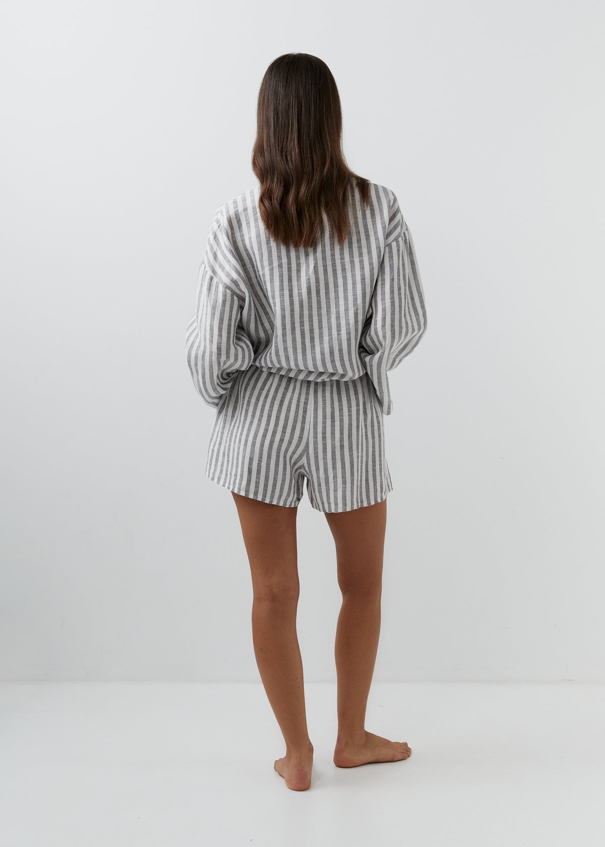 A woman viewed from behind wearing a matching gray and white vertically striped lounge set consisting of a long-sleeve top and shorts, standing barefoot against a plain white studio background.