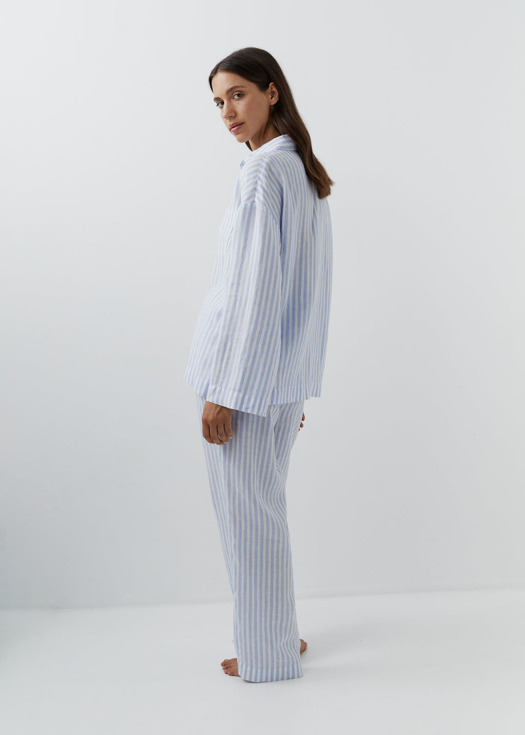 A woman modeling a matching two-piece pajama set with light blue and white vertical stripes, standing in profile against a plain white studio background.