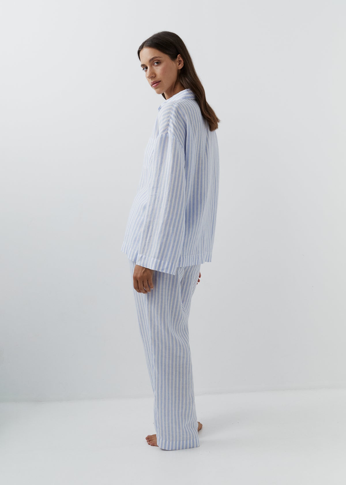 A woman modeling a matching two-piece pajama set with light blue and white vertical stripes, standing in profile against a plain white studio background.