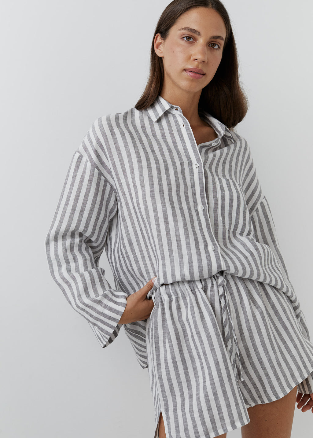 A woman wearing a loose, long-sleeve button-front shirt dress with vertical gray and white stripes, photographed against a plain light gray studio background.