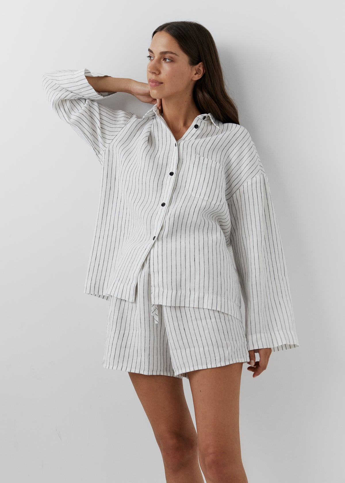 A woman modeling a matching two-piece pajama set consisting of a long-sleeve button-up shirt and shorts, both featuring a white base with thin vertical black pinstripes, set against a plain light gray wall.