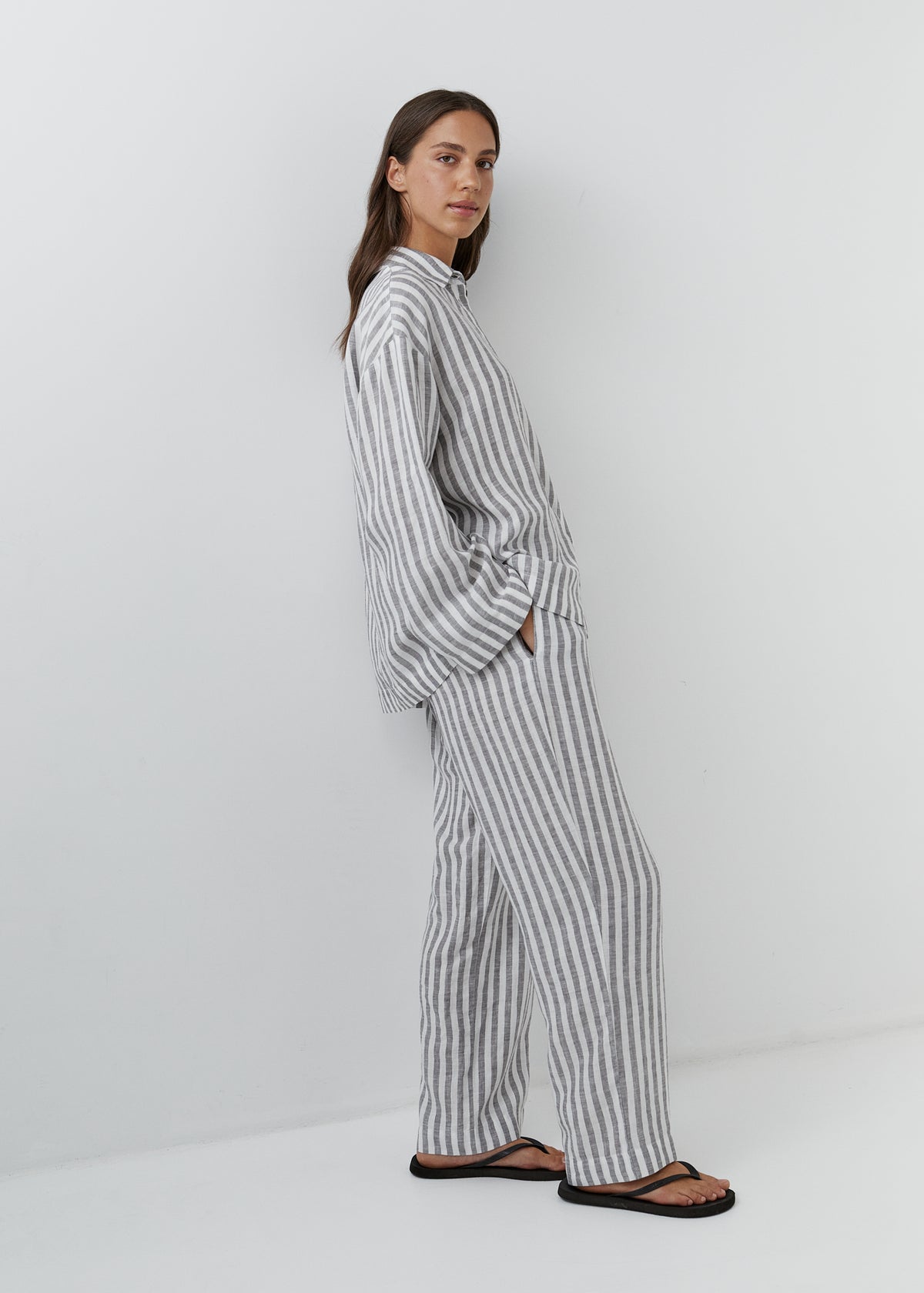A woman modeling a matching two-piece pajama set with vertical gray and white stripes, standing in a studio against a plain white background.