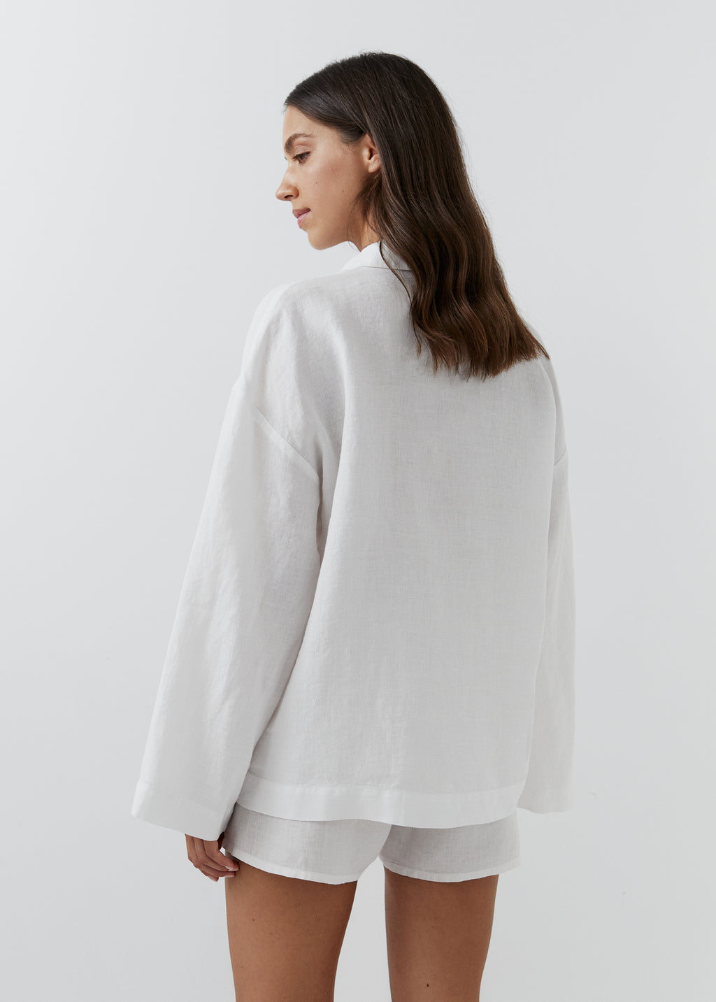 A woman shown from the back wearing a loose white long-sleeve top and matching white shorts against a plain light gray studio background.