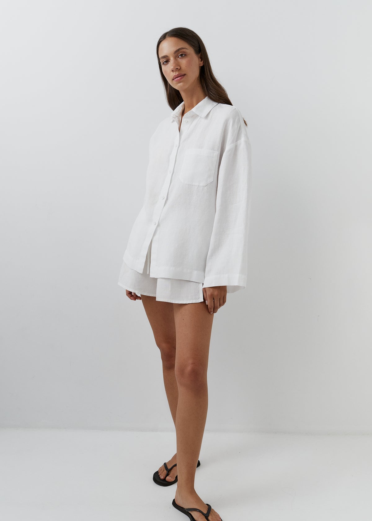A woman modeling a white linen button-up shirt and matching shorts set against a plain white studio background.