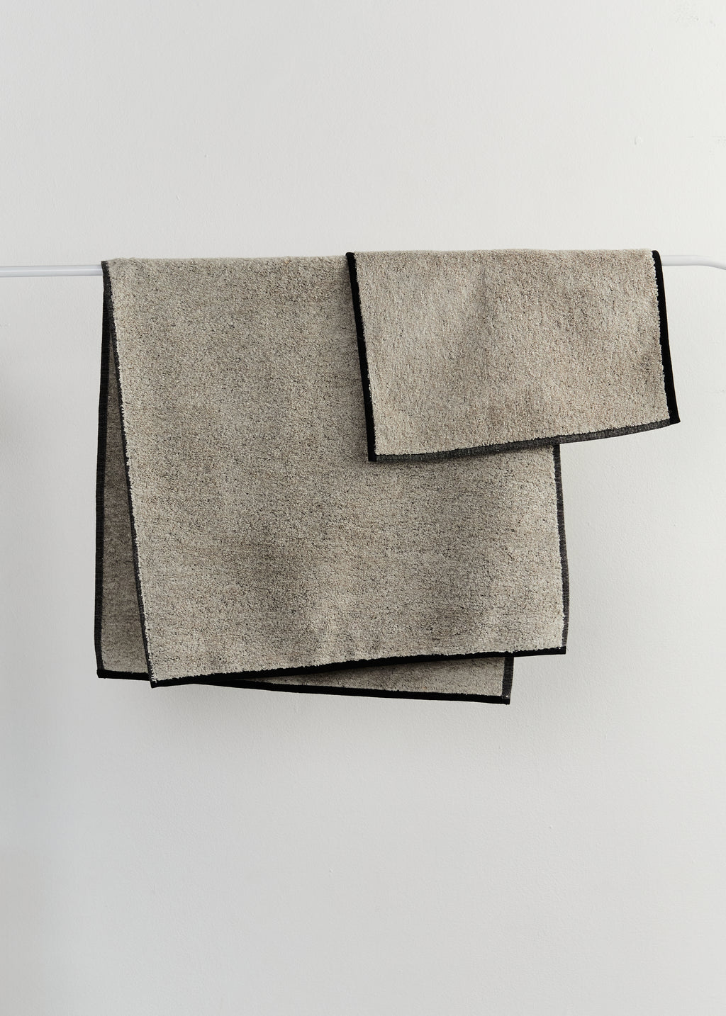 Two folded sand-coloured linen bath towels with contrasting black edges, hanging from a thin white rod against a light grey wall.
