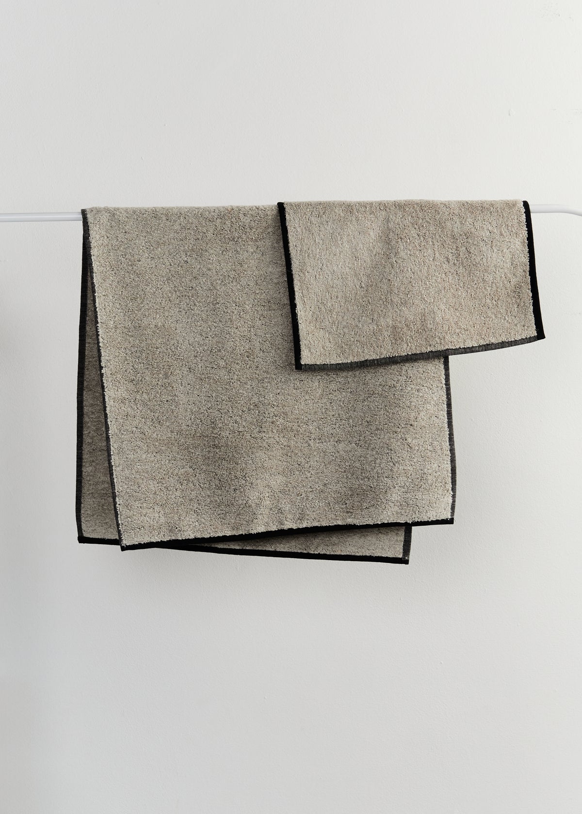 Two folded sand-coloured linen bath towels with contrasting black edges, hanging from a thin white rod against a light grey wall.

