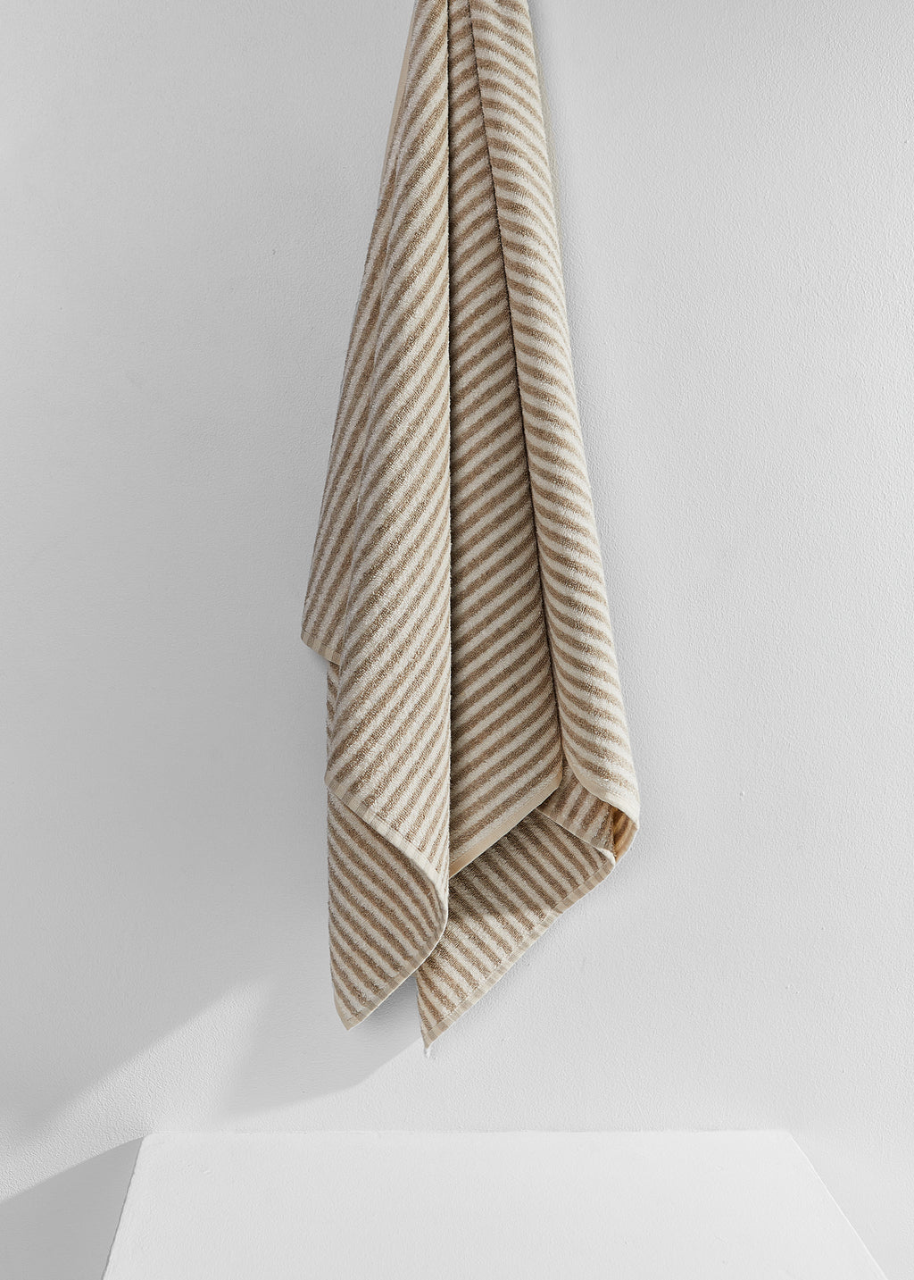 A beige and white striped linen bath towel hanging from a hook against a clean white wall.
