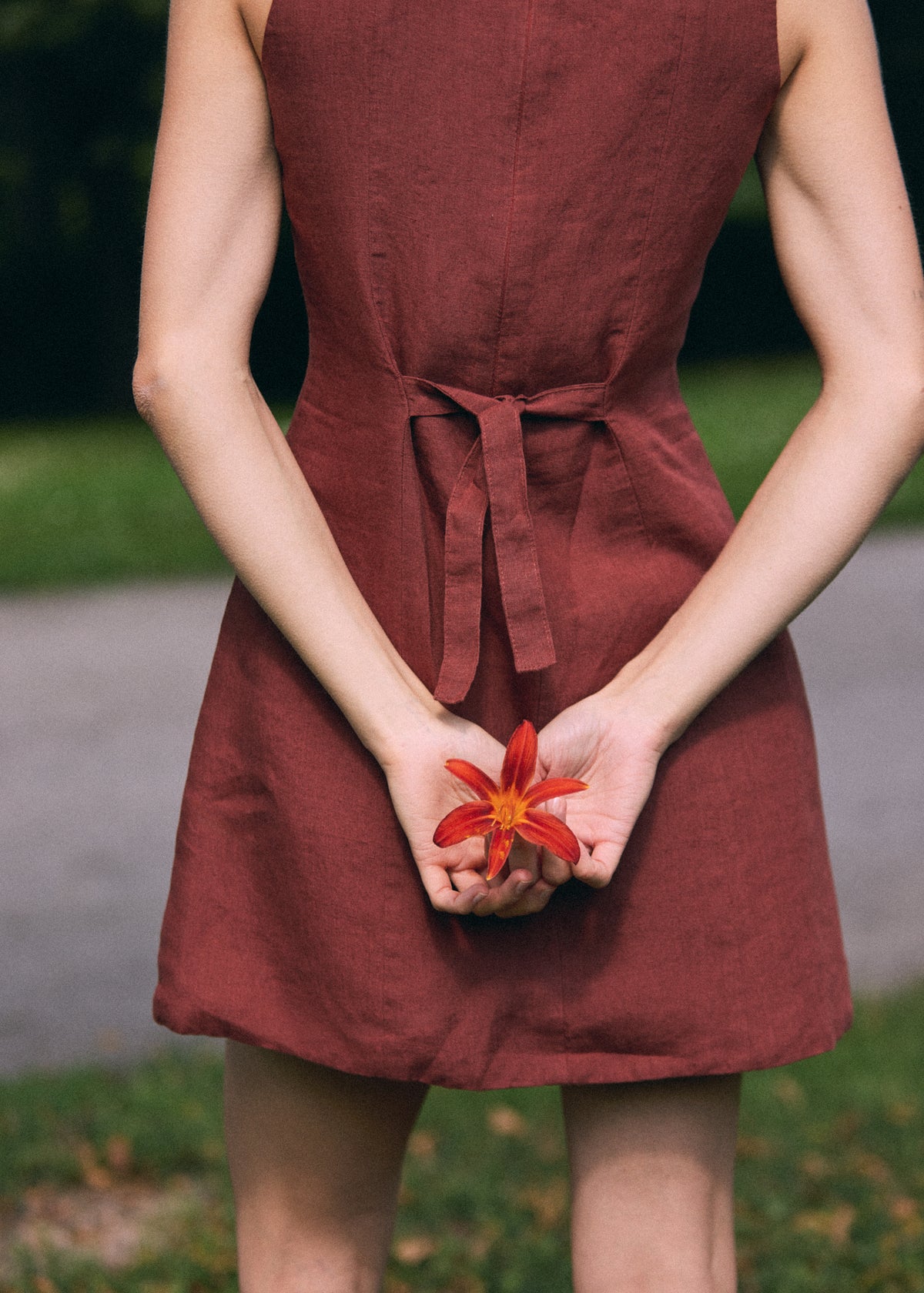 A person wearing a sleeveless rust-red linen dress, standing outdoors with their hands clasped in front holding a red flower.