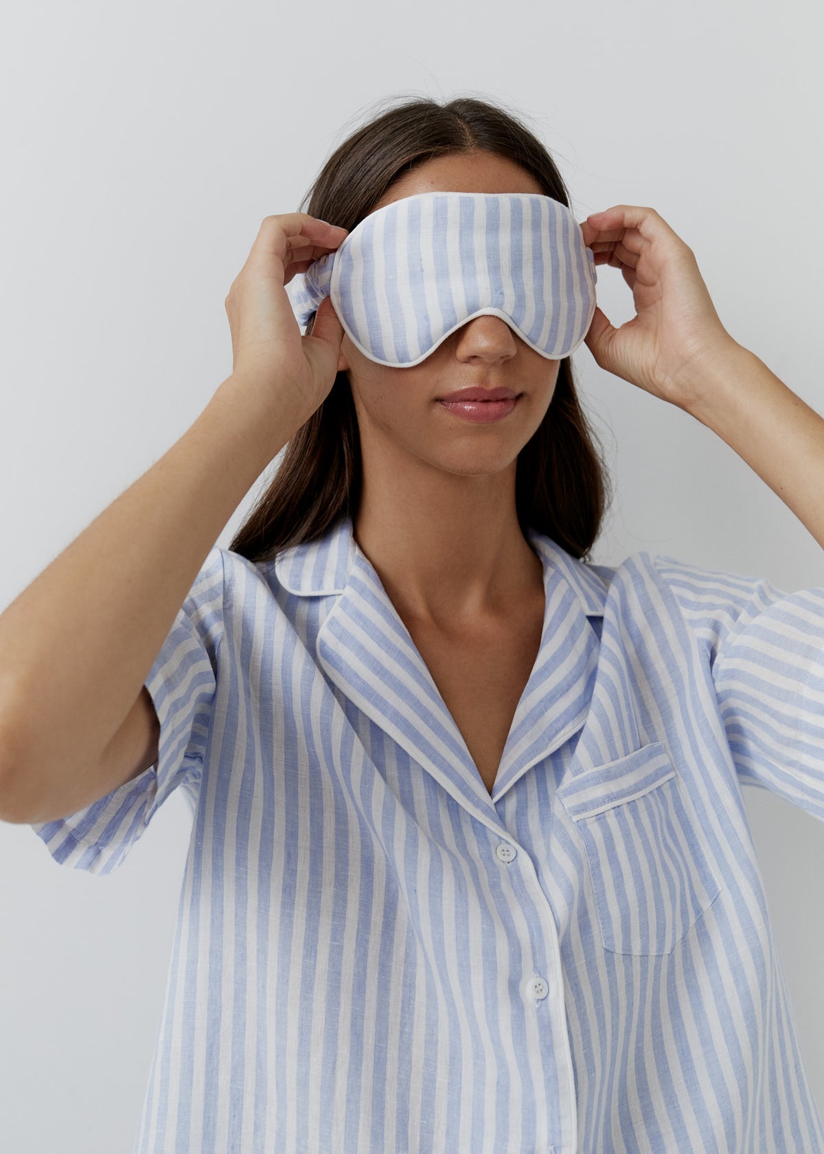 A woman wearing a light blue and white vertically striped sleep mask and matching pajama shirt, adjusting the strap of the mask against a plain light gray background.